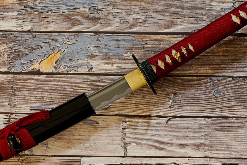 Katana tranchant, forge damassée, avec fourreau (saya 鞘) noire, tsuka-ito (柄糸) et sageo (下緒) rouge sombre, livré dans un coffret en bois contenant un kit d'entretien et une housse de protection en tissu | C’est dans un bain de sang que je suis née. Réputé mais discret, le forgeron qui m’a donné vie fut victime de son succès. Des brigands l’ont dépouillé alors même qu’il finissait de me forger. N’ayant alors pas de monture, ils ne prêtèrent pas attention à moi, posée sur le socle de polissage. Bien mal leur en prit, car lorsque le maître samouraï à qui j’étais destinée découvrit le forfait, il trouva un nouvel artisan pour créer ma monture, rouge sang.