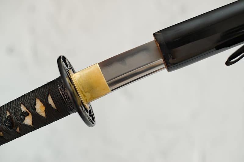 Katana tranchant, forge Kobuse (aciers 1045 / 1095), véritable ligne de trempe (hamon 刃文), affûtage à la main, avec fourreau (saya 鞘) noir, koiguchi (鯉口), Kurigata (栗形) et Kojiri (鐺) en corne naturelle, livré dans un coffret en bois composite contenant un kit d'entretien, un support laqué et une housse de protection en tissu | Portée par mon maître samouraï, j’ai sillonné la terre au point que ma nouvelle monture en porte la trace. Le cordage marron est désormais le symbole de mon nom, Seishi (聖地 Terre sacrée).