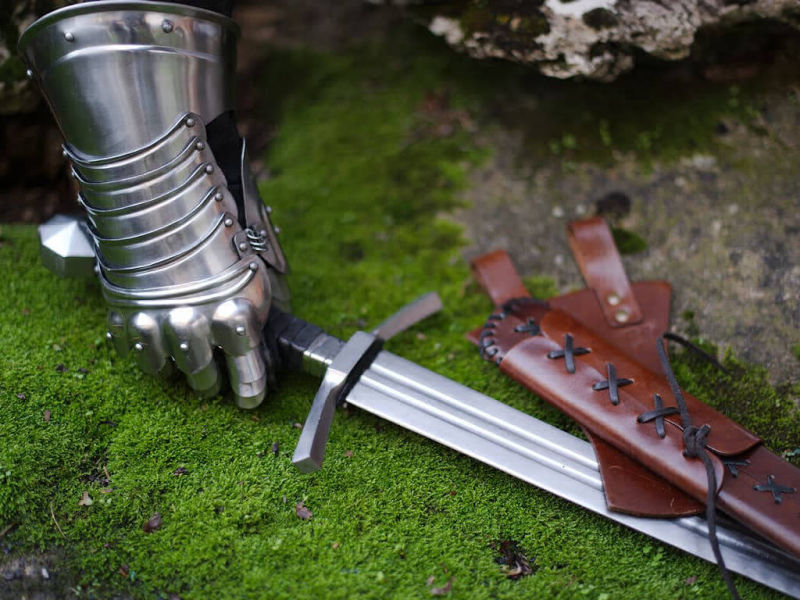 Une main de chevalier protégée par un gantelet articulé prend possession d'une épée médiévale forgée posée sur une roche recouverte de mousse