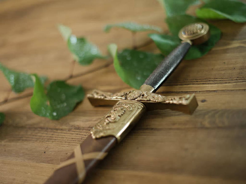 Excalibur protégée par son fourreau tressé, posée sur une table médiévale décorée de végétation et quelques gouttes de rosée