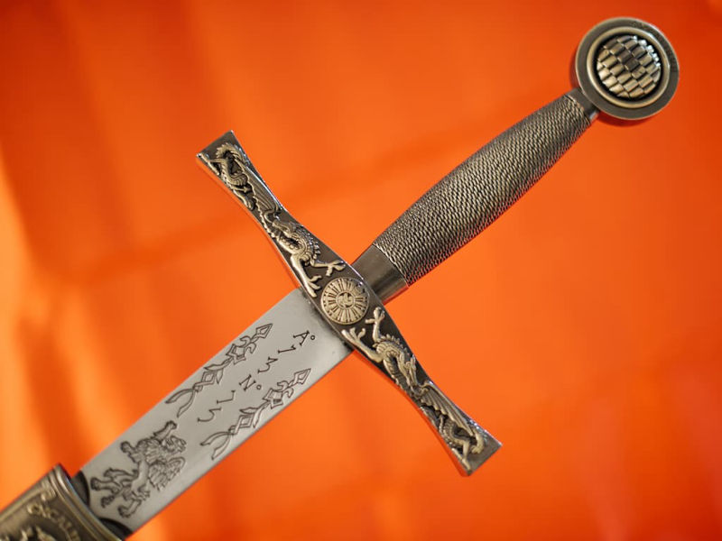 Excalibur protégée par son fourreau tressé, posée sur une table médiévale décorée de végétation et quelques gouttes de rosée Excalibur, l'Épée du roi Arthur, avec fourreau rouge tressé #Terressens