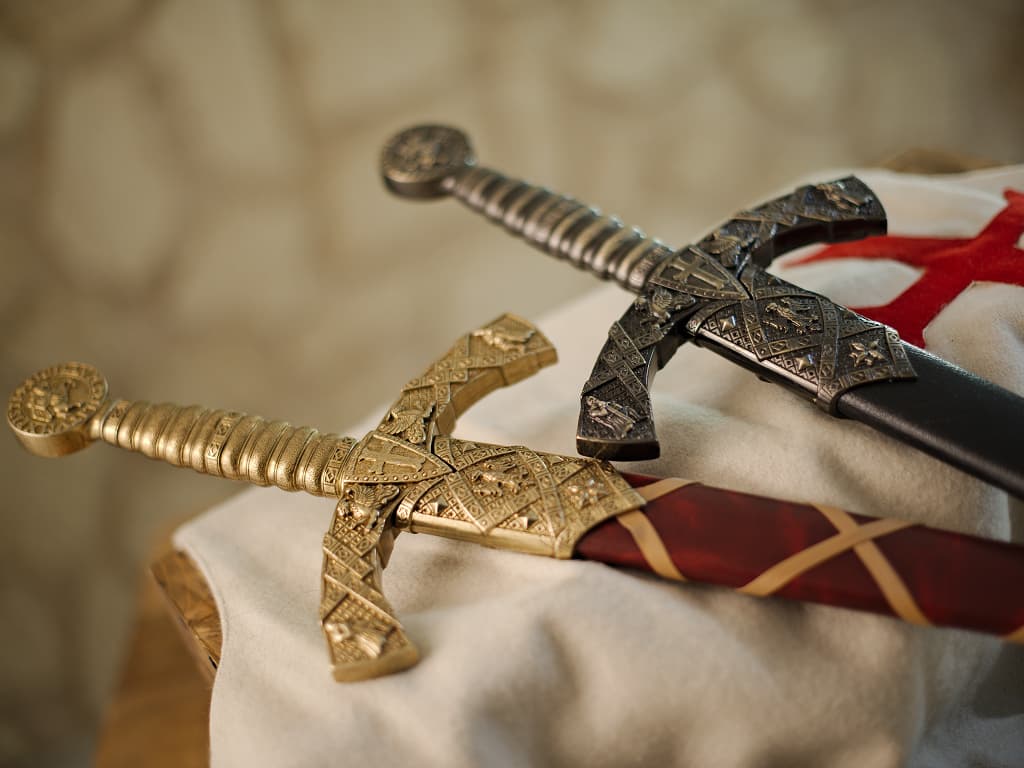 Les deux épées templières de décoration avec fourreau rouge tressé ou noir, posées sur un tabard templier blanc en laine, arborant la croix pattée rouge templière Les deux épées templières de décoration avec fourreau rouge tressé ou noir, posées sur un tabard templier blanc en laine, arborant la croix pattée rouge templière