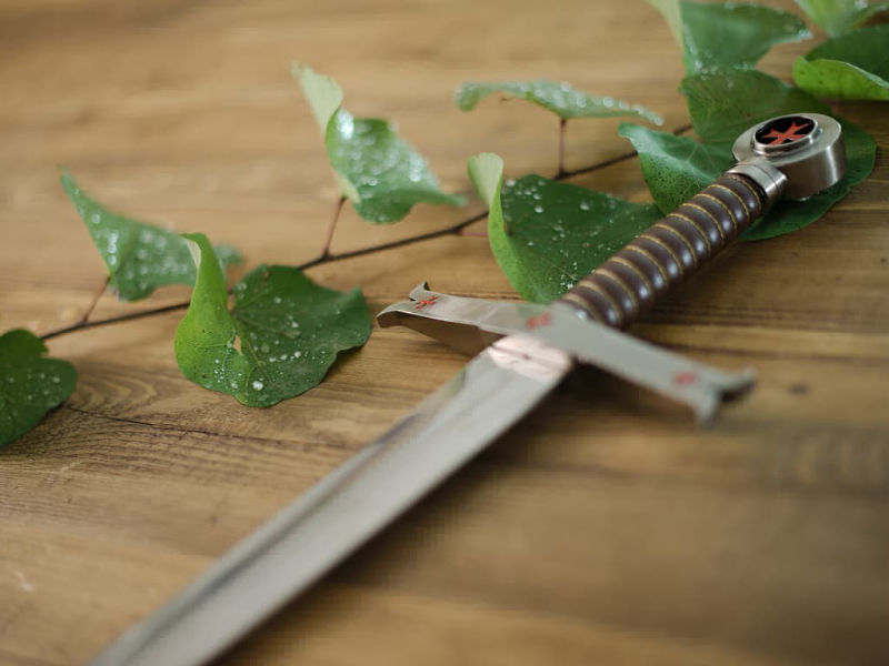 Épée templière de décoration posée sur une table médiébale en bois, avec une branche dont les branches abritent des gouttes de rosée Épée templière de décoration posée sur une table médiébale en bois, avec une branche dont les branches abritent des gouttes de rosée